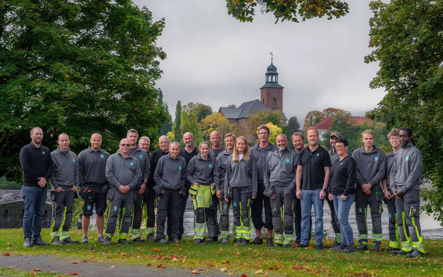 De ansatte i Ag, i grå arbeidsklær med Ag-logo, samlet på plenen ved Lågen foran Kongsberg kirke.